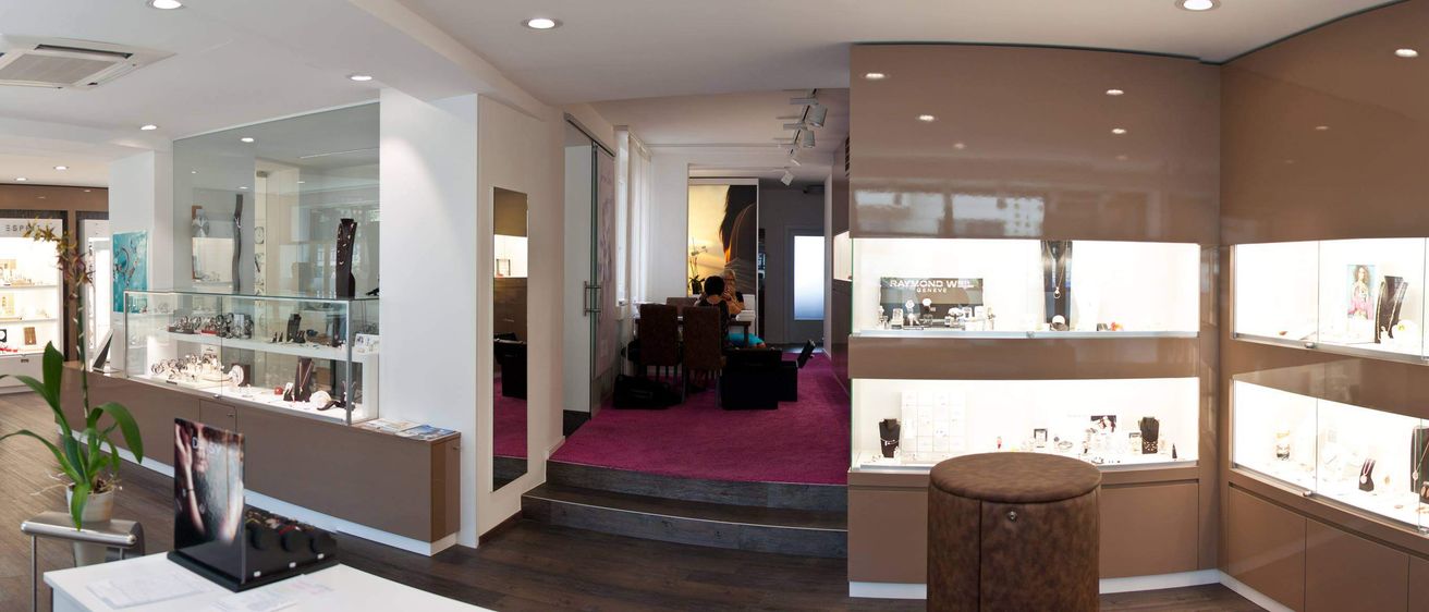 Ein Innenraum eines Geschäfts mit zwei Frauen auf einem Stuhl, einem Teppich und einem Tisch mit Stühlen, einem Spiegel und einer Glasvitrine.