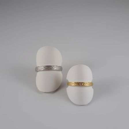 Zwei Ringe mit strukturierter Oberfläche, einer silber und einer golden mit einem kleinen Edelstein, auf weißen eiförmigen Objekten vor einem grauen Hintergrund präsentiert.