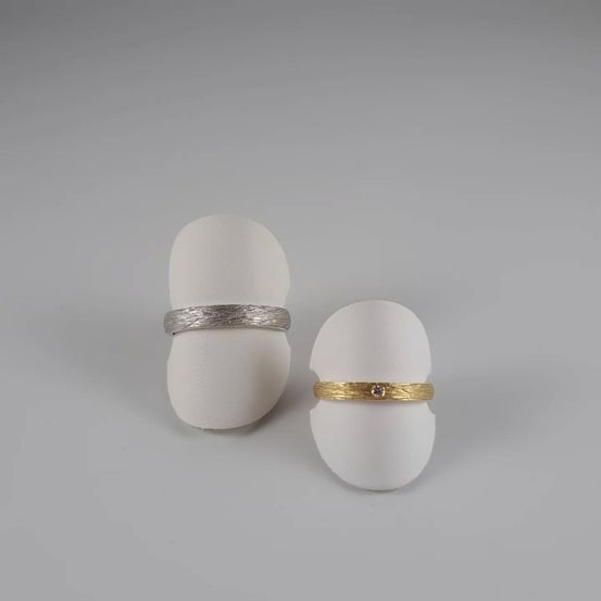 Zwei Ringe mit strukturierter Oberfläche, einer silber und einer golden mit einem kleinen Edelstein, auf weißen eiförmigen Objekten vor einem grauen Hintergrund präsentiert.