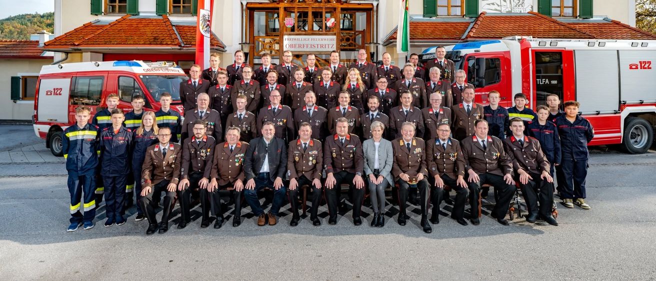 Eine Gruppe von Feuerwehrleuten und Beamten posiert für ein Foto vor einem Feuerwehrhaus mit Flaggen und einem Feuerwehrauto im Hintergrund.