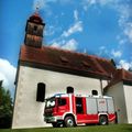 Ein roter und weißer Feuerwehrwagen steht vor einer Kirche mit einem roten Dach und einem Turm. Der Himmel ist blau mit einigen Wolken.