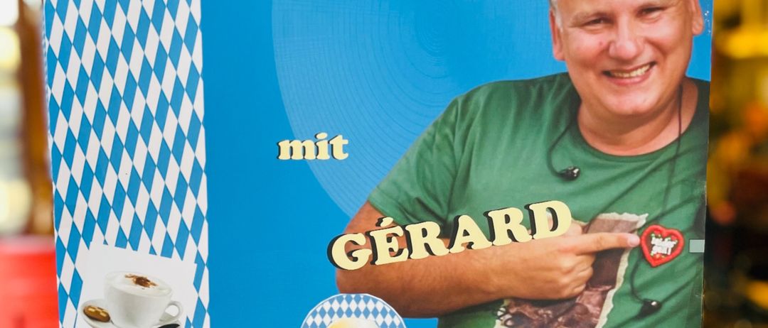 Ein Plakat für eine 'Fruischoppn mit Gerard' Veranstaltung im Cafe Harlekin am 9. August 2026, mit einem lächelnden Mann in einem grünen T-Shirt und einem Herz-Anstecker.