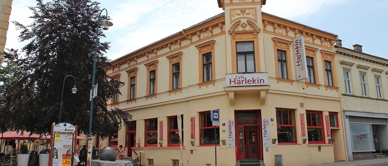 Cafe Harlekin, ein Gebäude mit einem Turm und mehreren Fenstern, befindet sich in einer Stadt mit klarem Himmel. Ein Schild zeigt den Namen und das Logo des Cafés an.