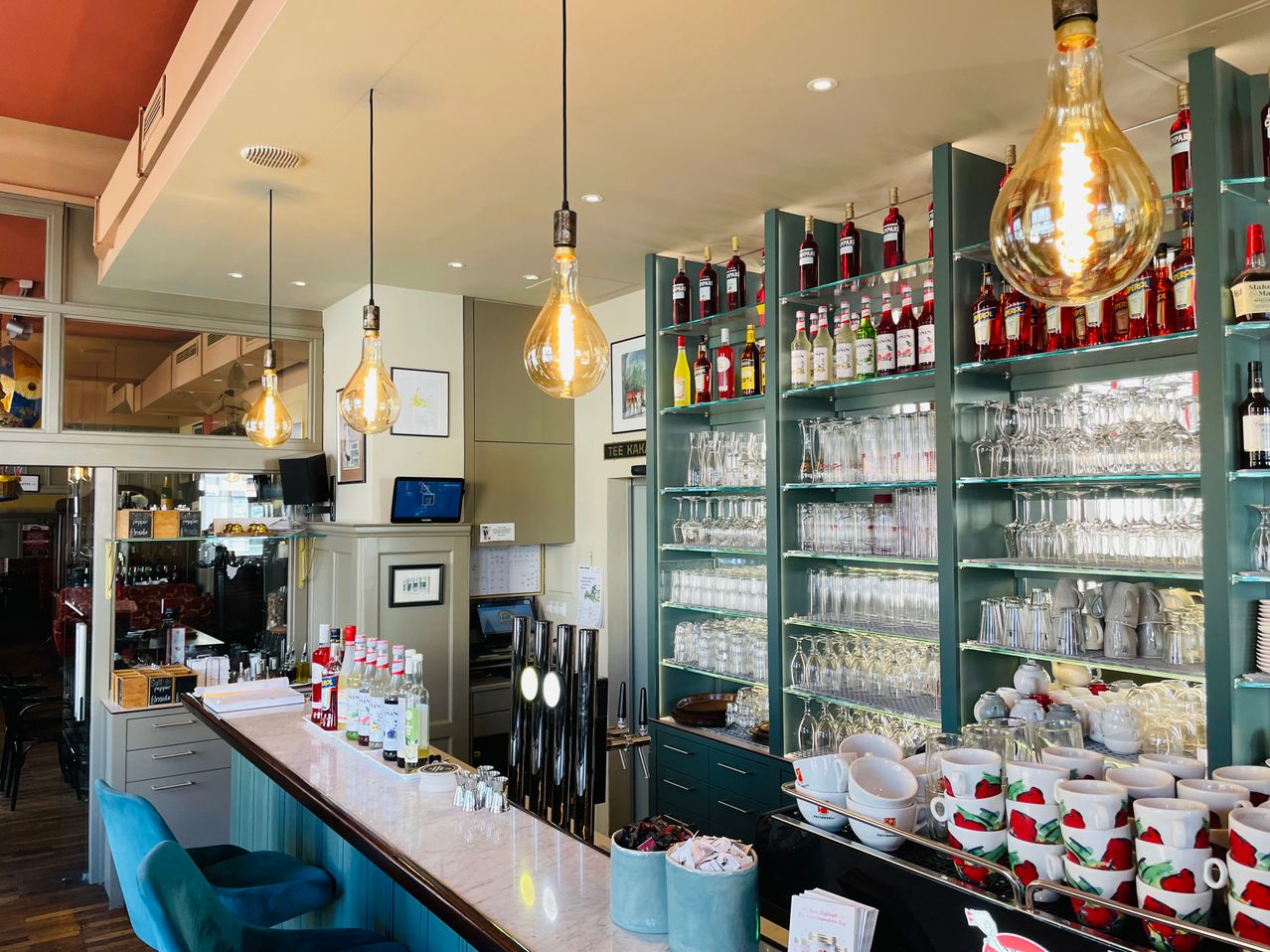 Eine Innenbar mit einer Marmortheke, blauen Stühlen und verschiedenen Flaschen und Gläsern in den Regalen. Die Decke hat Leuchtkörper und eine große Glühbirne.