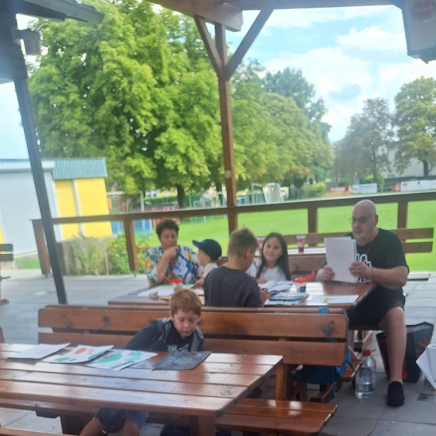 Eine Gruppe von Menschen sitzt an einem Picknicktisch im Freien, ein Mann liest eine Zeitung. Im Hintergrund sind Bäume und ein Rasenfeld zu sehen.