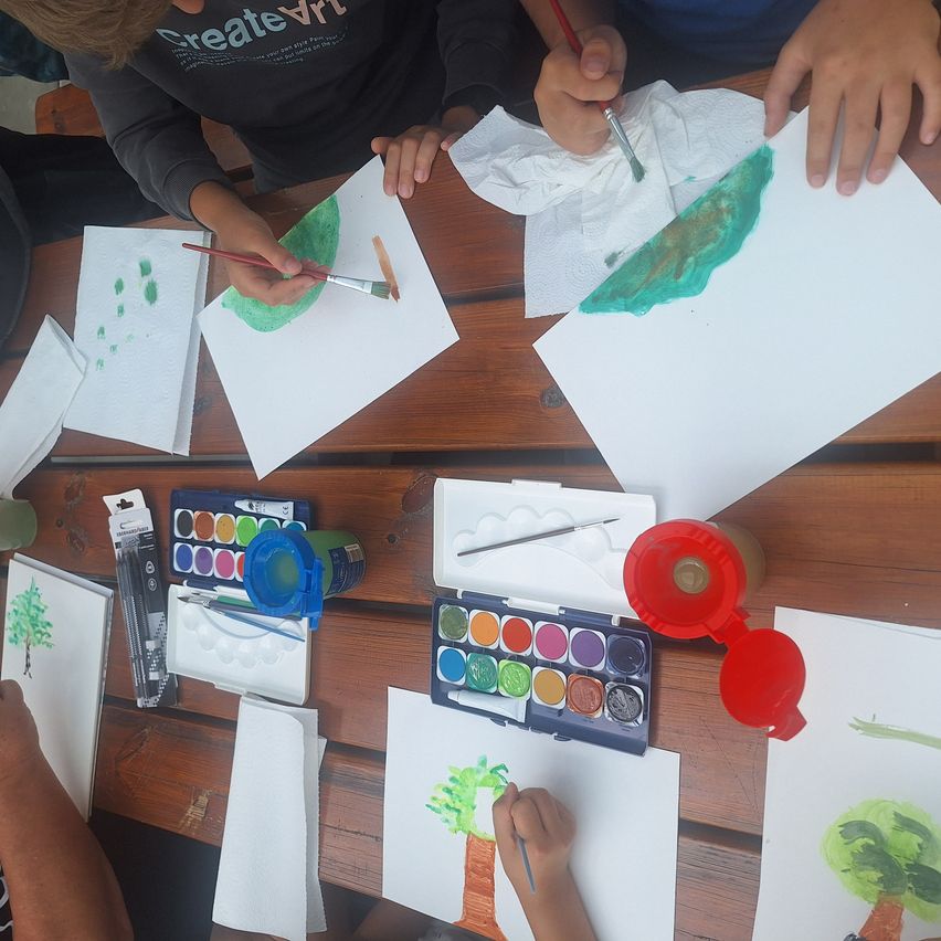 Zwei Kinder malen auf weißen Papieren mit Pinsel. Ein Kind malt ein grünes Blatt, während das andere einen Baum malt. Aquarellsets und Flaschen sind auf dem Tisch.