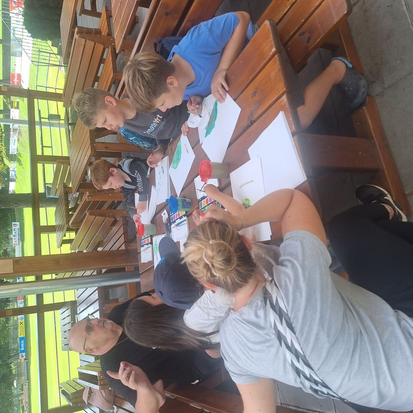Vier Kinder malen an einem Tisch mit Erwachsenen in der Nähe, die auf hölzernen Bänken sitzen. Der Tisch hat Malwerkzeuge und Papier. Die Kinder konzentrieren sich auf ihre Kunstwerke.