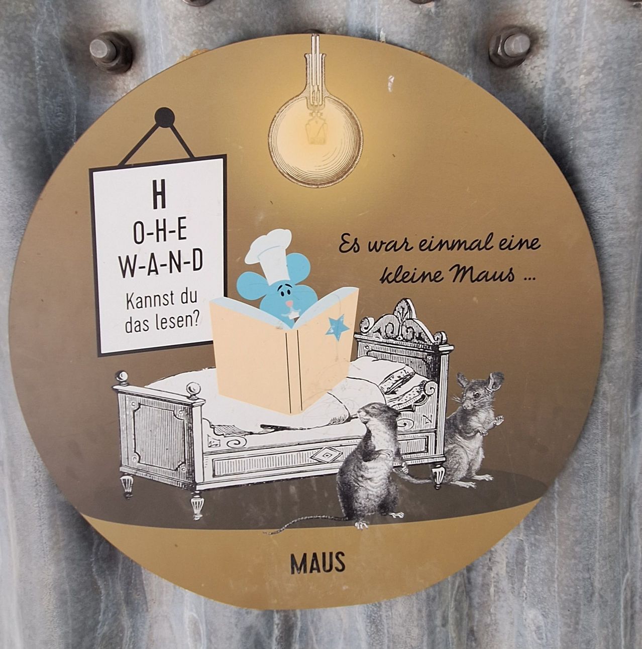 Ein Schild mit einer Maus, die ein Buch liest, darunter steht 'Es war einmal eine kleine Maus...' und 'Kannst du das lesen?' Es ist an einer Metalloberfläche befestigt.