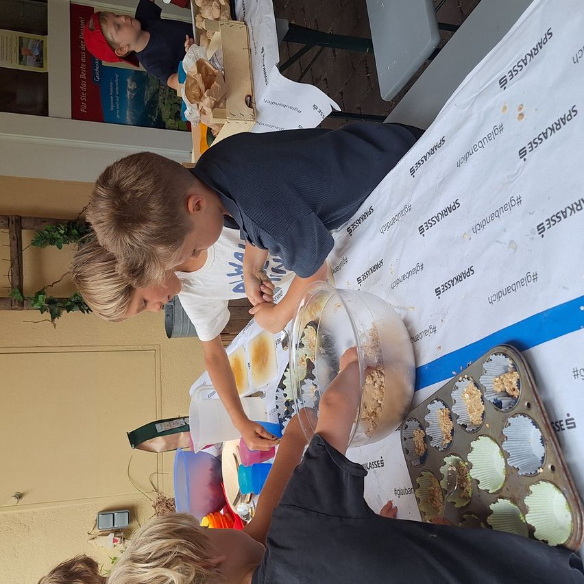 Mehrere Kinder versammeln sich um einen Tisch und backen. Sie tragen Freizeitkleidung und scheinen Spaß zu haben.