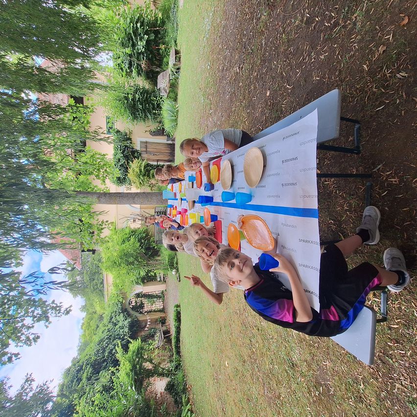 Kinder versammeln sich um einen Tisch im Freien mit einem Banner und genießen eine Mahlzeit unter Bäumen. Sie tragen Freizeitkleidung und scheinen für ein Foto zu posieren.