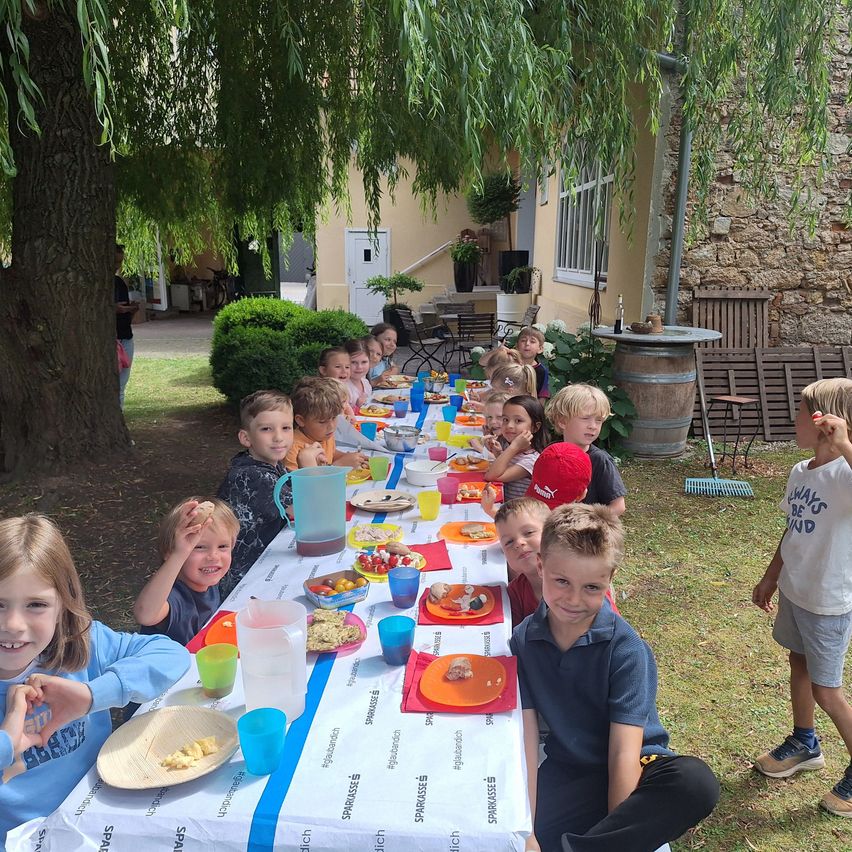 Eine Gruppe von Kindern sitzt um einen langen Tisch im Freien, mit Essen, Bechern und Tellern. Einige tragen Mützen. Der Tisch ist mit einem weißen Tuch bedeckt. Ein Baum steht in der Nähe und ein Gebäude mit einer Steinmauer im Hintergrund.