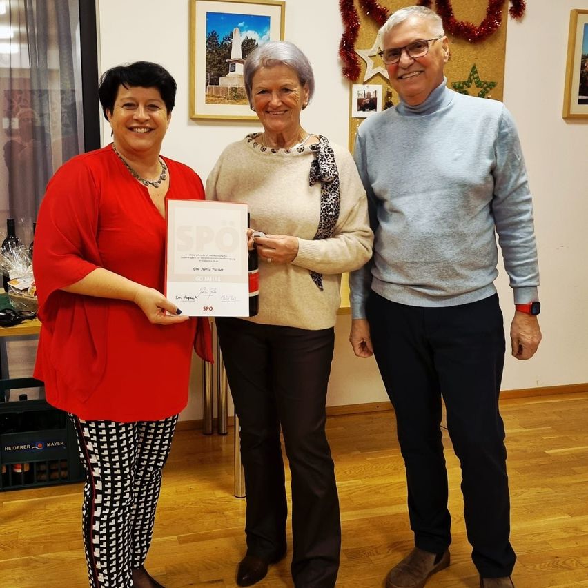 Drei Personen stehen in einem Raum, eine hält ein Zertifikat mit der Aufschrift SPD. Die Frau auf der linken Seite trägt eine rote Bluse und gemusterte Hose. Die Frau in der Mitte hat ein Tuch und trägt einen beigen Pullover. Der Mann auf der rechten Seite trägt ein blaues Rollkragenhemd und schwarze Hose.