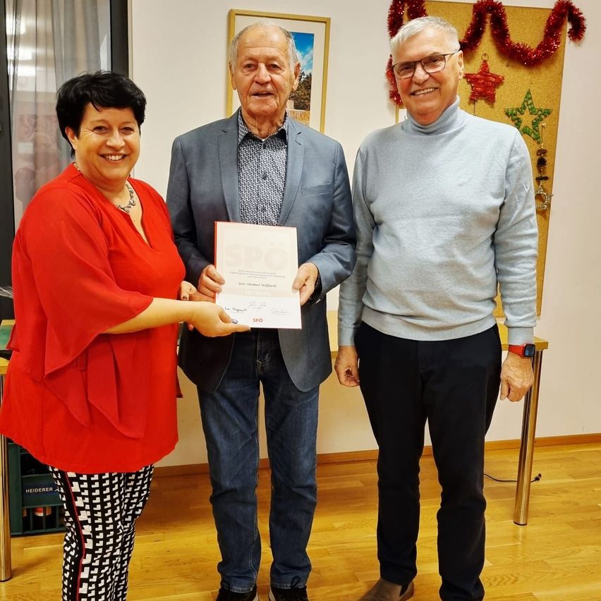 Eine Frau und zwei Männer stehen in einem Raum. Der Mann in der Mitte hält ein Zertifikat, und die Frau reicht es ihm. Sie alle lächeln. Hinter ihnen ist eine Wand mit Dekorationen.