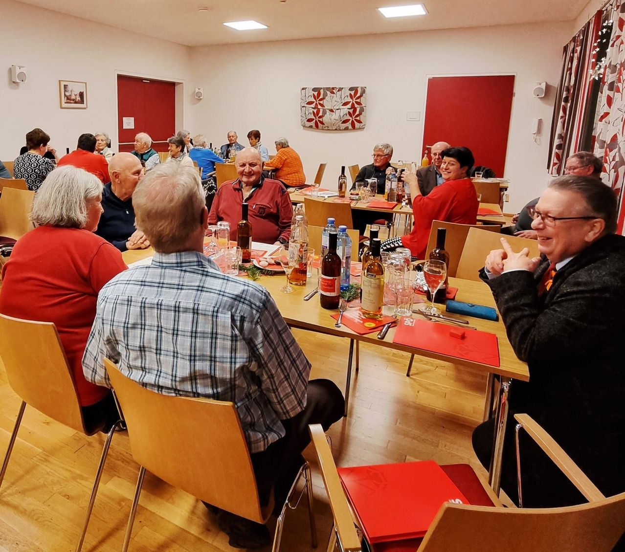 Eine Gruppe älterer Menschen sitzt um einen Esstisch herum und genießt eine Mahlzeit mit Getränken. Der Tisch ist mit Flaschen, Gläsern und Tellern gedeckt. Der Raum hat weiße Wände und rote Türen.