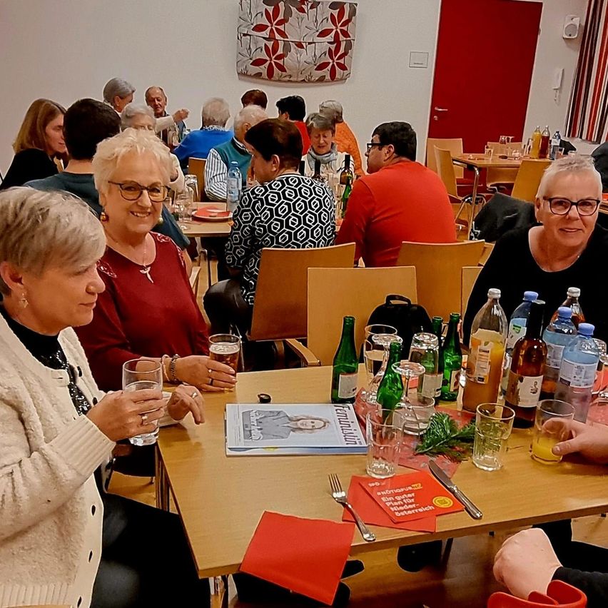 Eine Gruppe von Menschen sitzt an einem Tisch in einem Restaurant. Flaschen, Gläser und ein Buch sind auf dem Tisch. Einige Leute tragen Brillen.