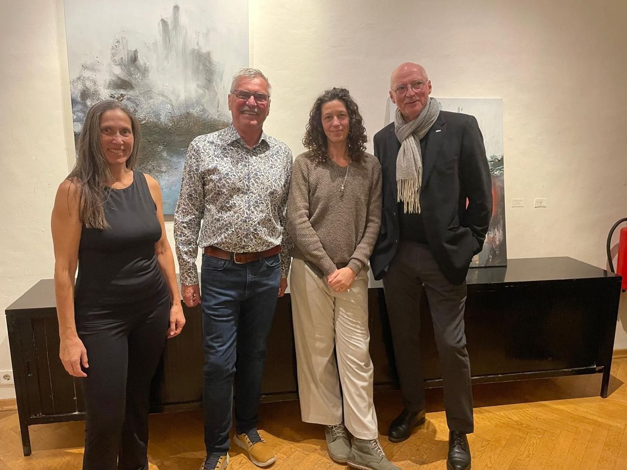 Vier Personen, darunter zwei Männer und zwei Frauen, stehen zusammen vor einer Wand mit abstrakten Gemälden und lächeln und posieren für ein Foto.