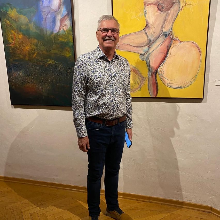 Ein älterer Mann mit Brille steht in einer Kunstgalerie, lächelt und hält ein Telefon. Hinter ihm befinden sich zwei Gemälde, eines mit einer Figur auf gelbem Hintergrund und ein weiteres mit einer farbenfrohen abstrakten Landschaft.
