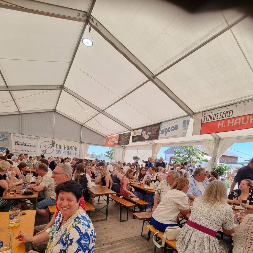 Ein großes Zelt mit vielen Menschen an Tischen, die Essen und Getränke genießen. Banner hängen an der Decke, und einer lautet Schlosserei.