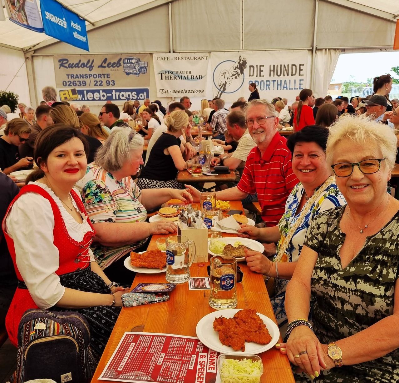 Gruppe von Menschen sitzt an einem Tisch in einem Restaurant. Zwei Frauen und zwei Männer, lächelnd zur Kamera. Eine Speisekarte und Essen auf dem Tisch. Eine Frau trägt ein traditionelles Outfit.