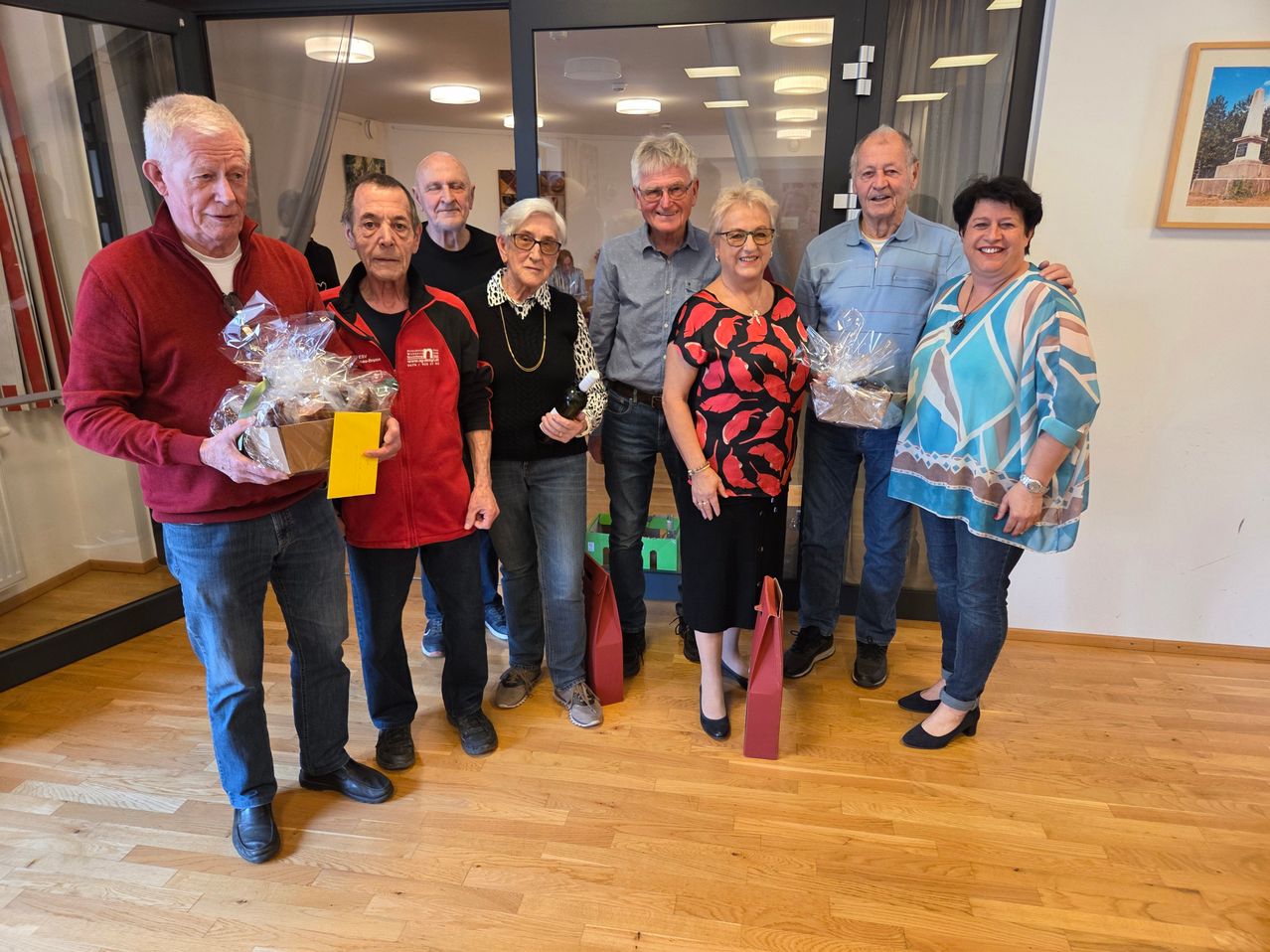 A group of elderly individuals are posing for a photograph, smiling and holding gift baskets. They are standing on a wooden floor.