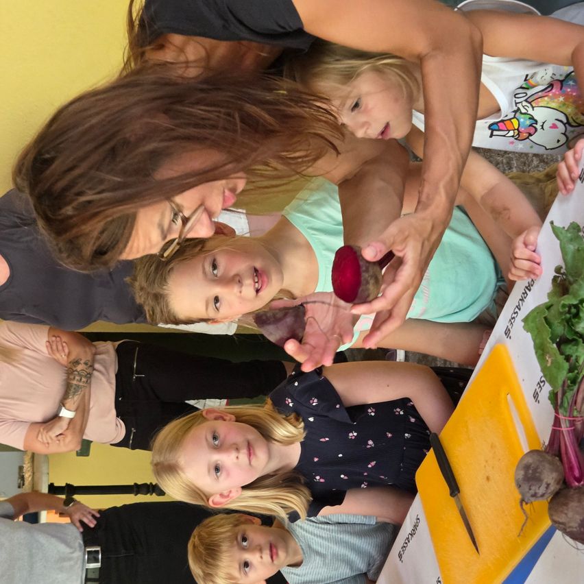 A group of children and adults are gathered around a table with vegetables, likely in a cooking class. The woman in glasses is holding a beetroot.