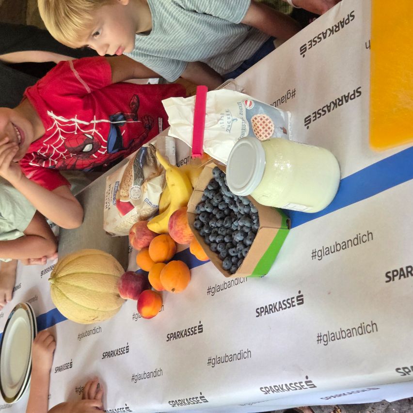 A table with various fruits, yogurt, and other food items. A child in a red shirt is looking at the camera.
