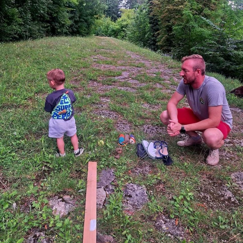 A man kneels on a grassy path, smiling, with a young boy standing nearby. Shoes, a stick, and a small toy are on the ground.