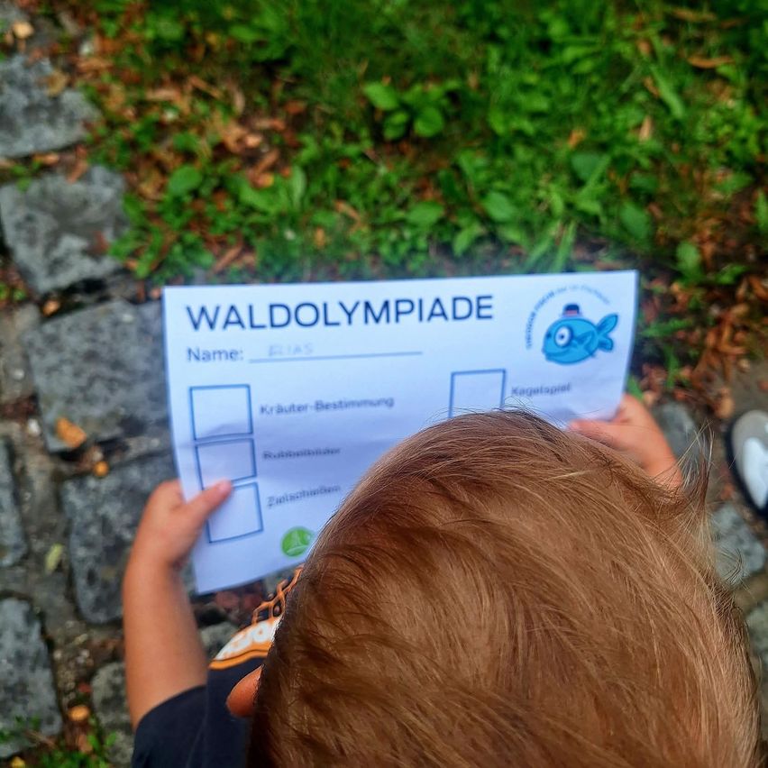A young child holds a paper titled 'WALDOLYMPIADE' with a fish logo, possibly participating in a nature-themed activity.
