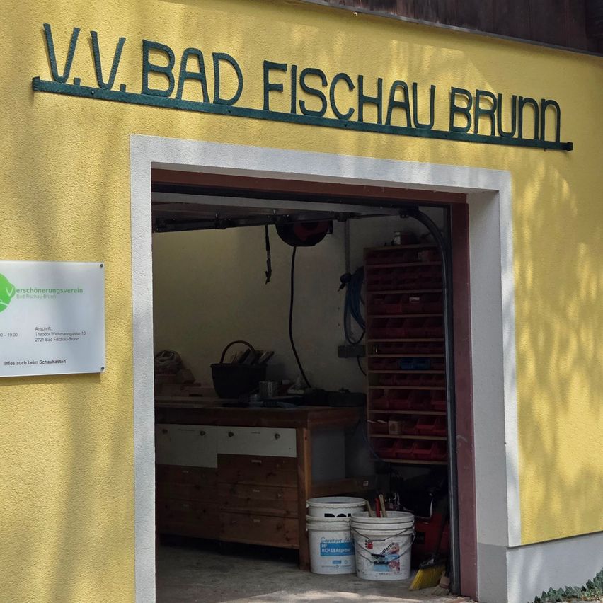 Ein gelbes Gebäude mit einem Schild, das V.V. Bad Fischau Brunn liest. Der Eingang ist offen und zeigt einen Holzschreibtisch und Regale im Inneren. Zwei Eimer und ein Besen sind in der Nähe des Eingangs zu sehen.