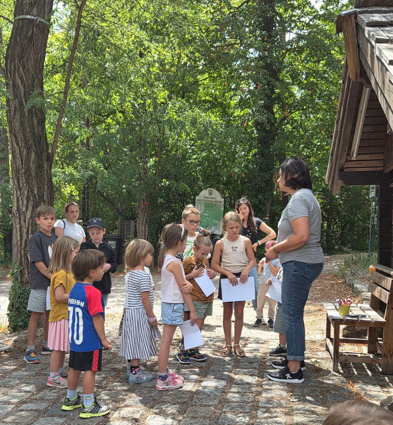 Eine Gruppe von Kindern und Erwachsenen versammelt sich in einem Park und hört einer Frau zu, die ein Stück Papier in der Hand hält. Die Kinder halten Papiere in der Hand und einige schauen darauf. Im Hintergrund befinden sich eine Holzstruktur und ein Schild.