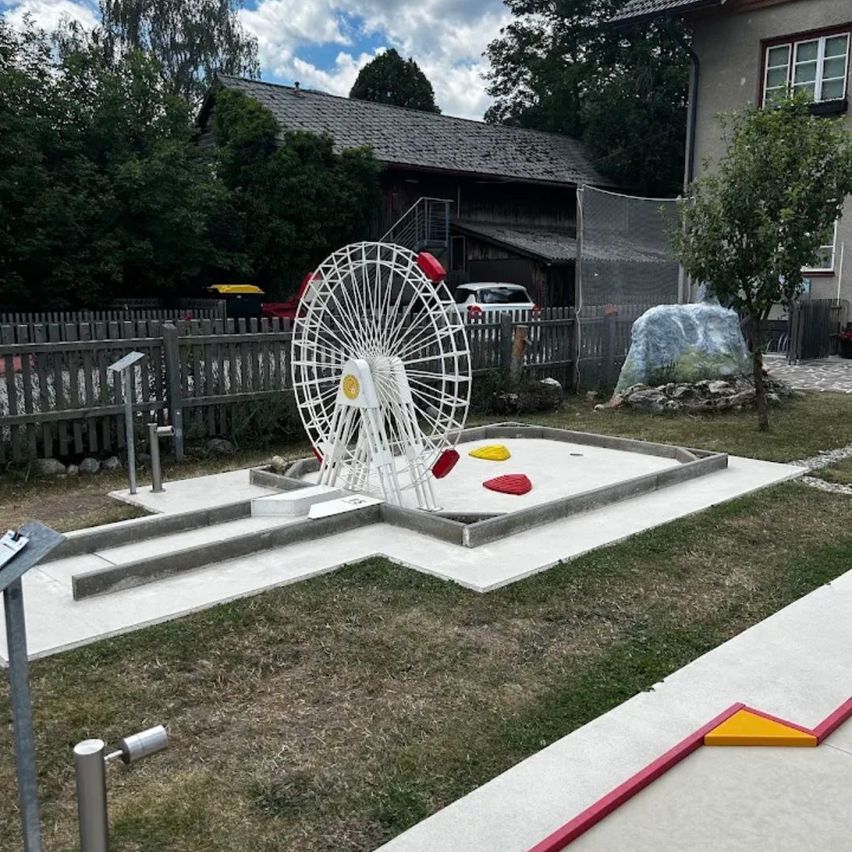 Ein Minigolfplatz mit einem Karussell in der Mitte, umgeben von einem Holzzaun und einem Haus im Hintergrund.
