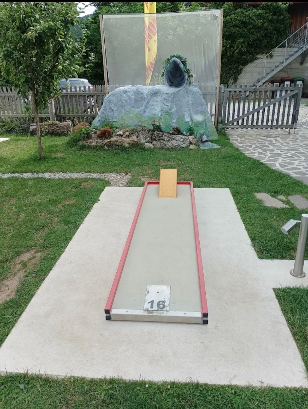 Ein Minigolfplatz mit einem roten und weißen Abschlagfeld, das mit 16 beschriftet ist, umgeben von einem Rasenbereich und einer großen Felsskulptur im Hintergrund.