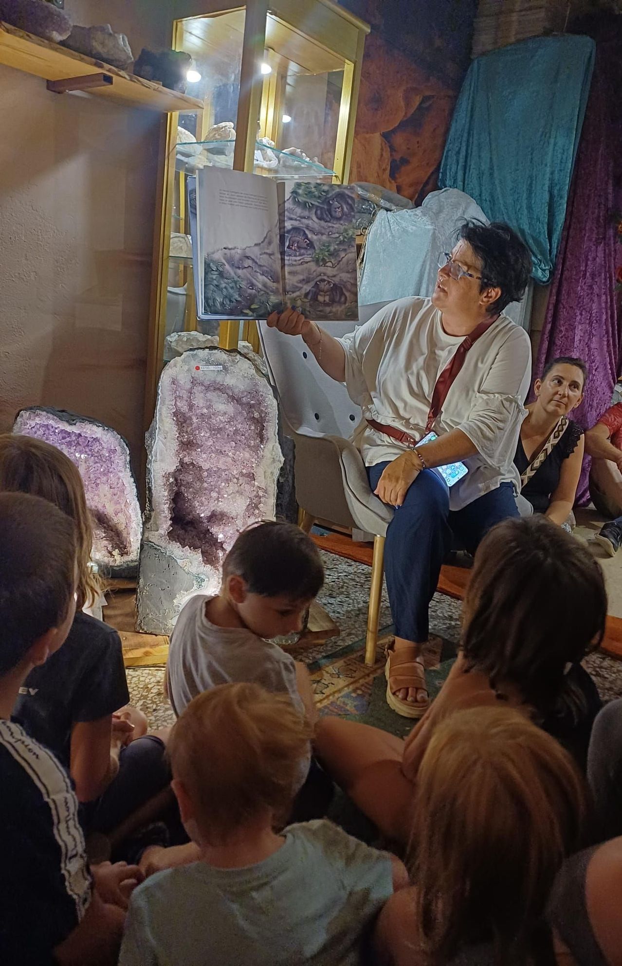 Eine Frau liest ein Buch vor einer Gruppe von Kindern, die im Kreis sitzen, mit großen Amethystkristallen im Hintergrund.