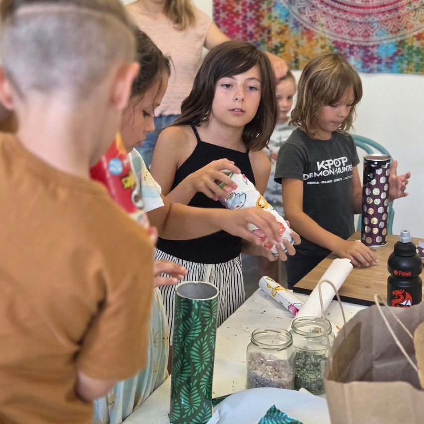 Eine Gruppe von Kindern ist um einen Tisch versammelt und beschäftigt sich mit einer Aktivität. Ein Kind hält ein eingewickeltes Geschenk, während andere zusehen. Der Tisch ist mit verschiedenen Gegenständen übersät, darunter Gläser, eine Tasche und ein gemusterter Becher.