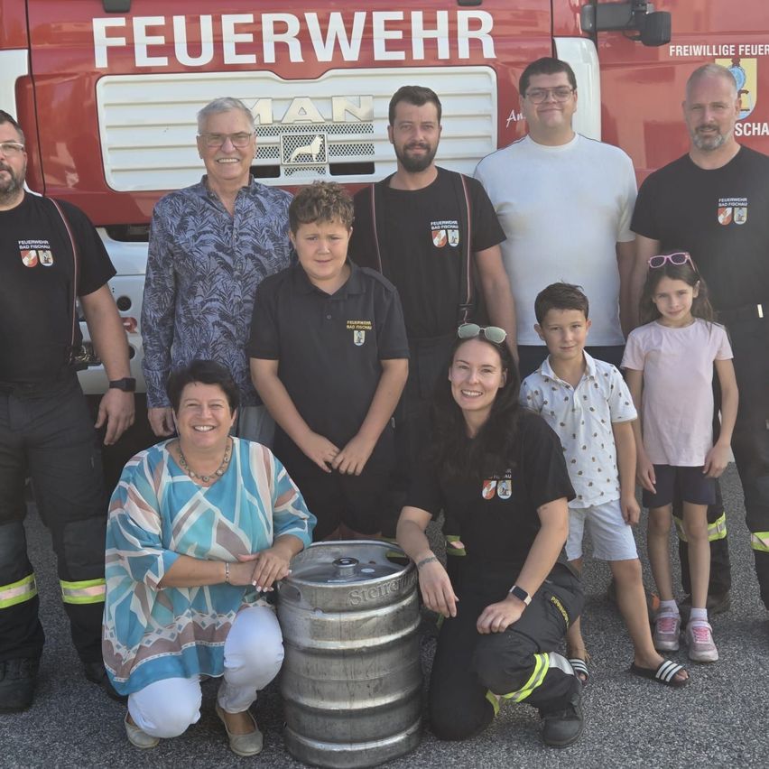 Eine Gruppe von Feuerwehrleuten und ihren Familien posiert für ein Foto vor einem Feuerwehrauto. Die Feuerwehrleute tragen Uniformen und einige halten einen großen Bierfass.