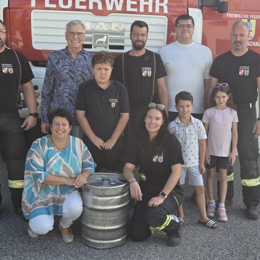Eine Gruppe von Menschen, darunter Feuerwehrleute und Kinder, posiert für ein Foto vor einem Feuerwehrauto. Sie lächeln und scheinen eine gute Zeit zu haben.