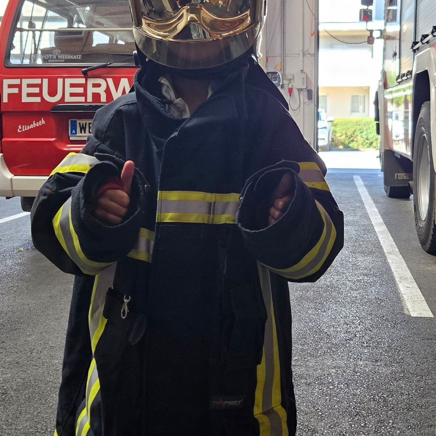 Ein junges Kind in einer Feuerwehruniform, das einen Daumen hochhält, steht vor einem roten Feuerwehrauto.