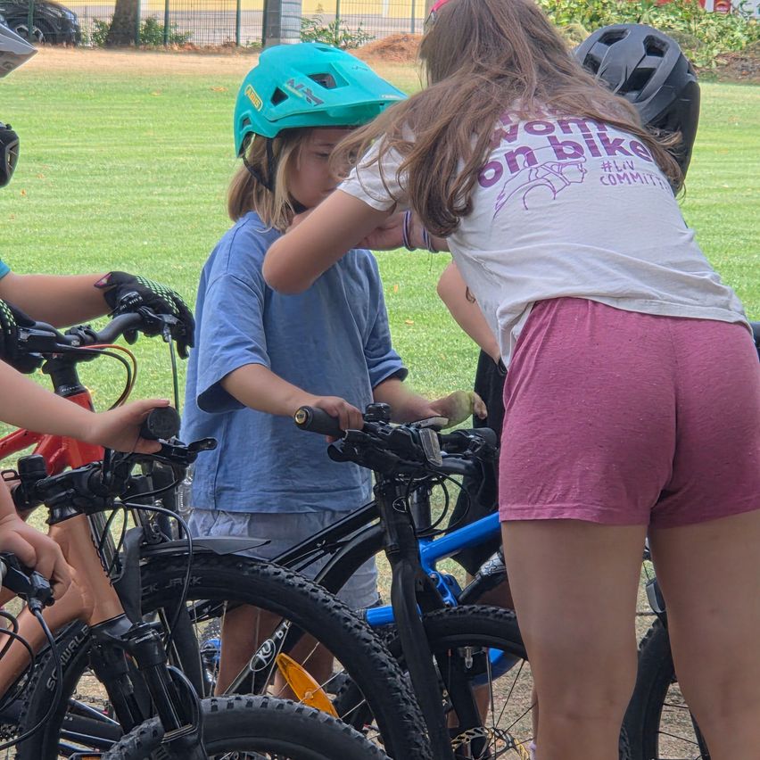 Eine Gruppe von Kindern versammelt sich in einem Park um Fahrräder. Sie tragen Helme und scheinen bereit zu sein, zu fahren. Das Mädchen vorne trägt ein weißes Shirt mit lila Text.