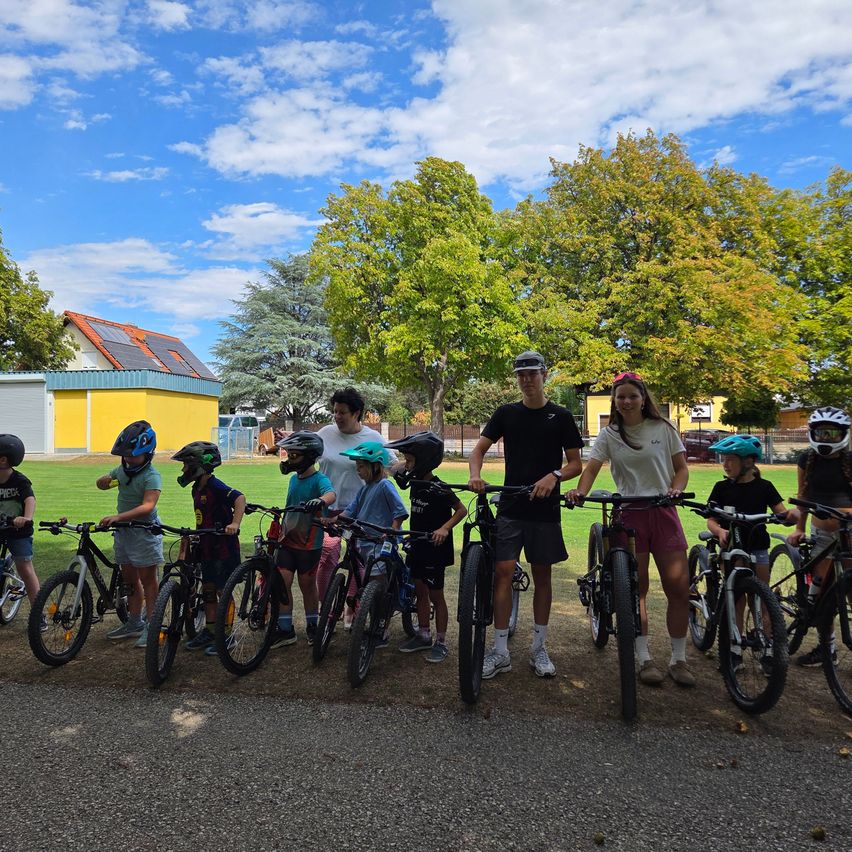 Eine Gruppe von Kindern und Erwachsenen steht mit ihren Fahrrädern in einem Park an einem sonnigen Tag. Die Kinder tragen Helme und einige Erwachsene tragen Brillen. Bäume und ein Gebäude mit Solarpanelen sind im Hintergrund zu sehen.