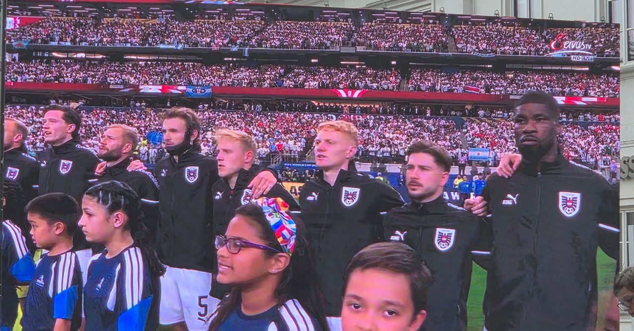 Eine Gruppe von Athleten steht vor einer jubelnden Menge in einem Stadion. Ein junges Mädchen und ein Junge, mit Brille und Stirnband, lächeln in die Kamera. Die Athleten tragen schwarze Uniformen.