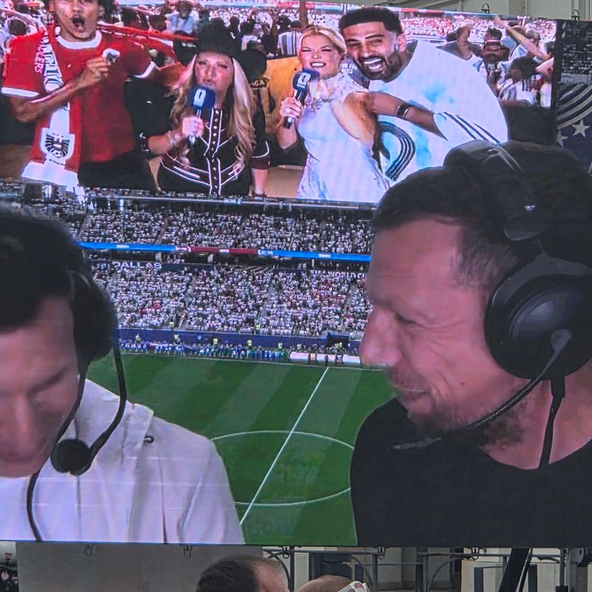 Zwei Kommentatoren mit Mikrofonen, einer in einem roten Shirt, der andere in einem weißen Shirt, interviewen ein Paar vor einer großen Menge. Ein Stadion mit einem Feld und einer großen Leinwand im Hintergrund.