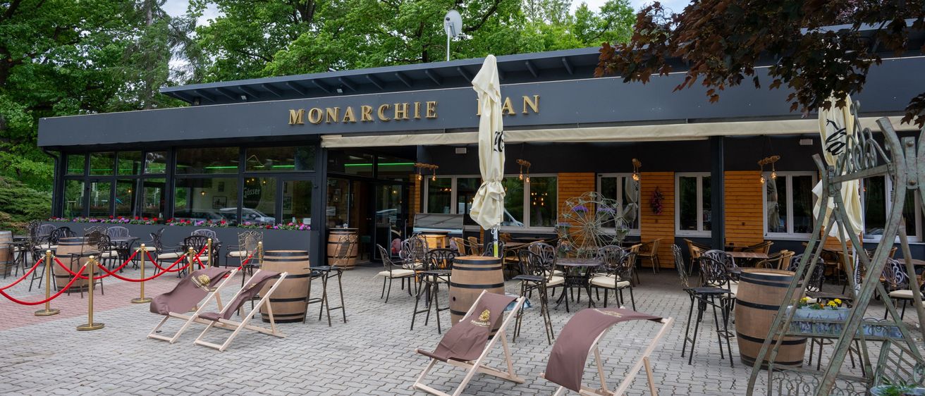 Im Freien Sitzbereich des Restaurants 'Monarchie' mit Holzfässern, Tischen, Stühlen und einem Schirm, umgeben von Grün.