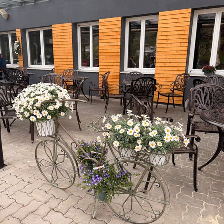 Ein dekoratives Fahrrad mit Blumen steht vor einem Gebäude mit Holzverkleidungen und Glasfenstern. Die Umgebung umfasst Tische und Stühle.