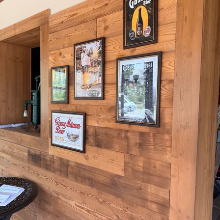 Eine Holzwand mit mehreren gerahmten Bildern und Schildern, eines davon sagt 'Gösser Maizen-Bier'. Darunter befindet sich ein Tisch mit Papieren darauf.