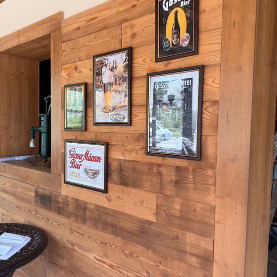 Eine Holzwand mit mehreren gerahmten Bildern und Schildern, eines davon sagt 'Gösser Maizen-Bier'. Darunter befindet sich ein Tisch mit Papieren darauf.