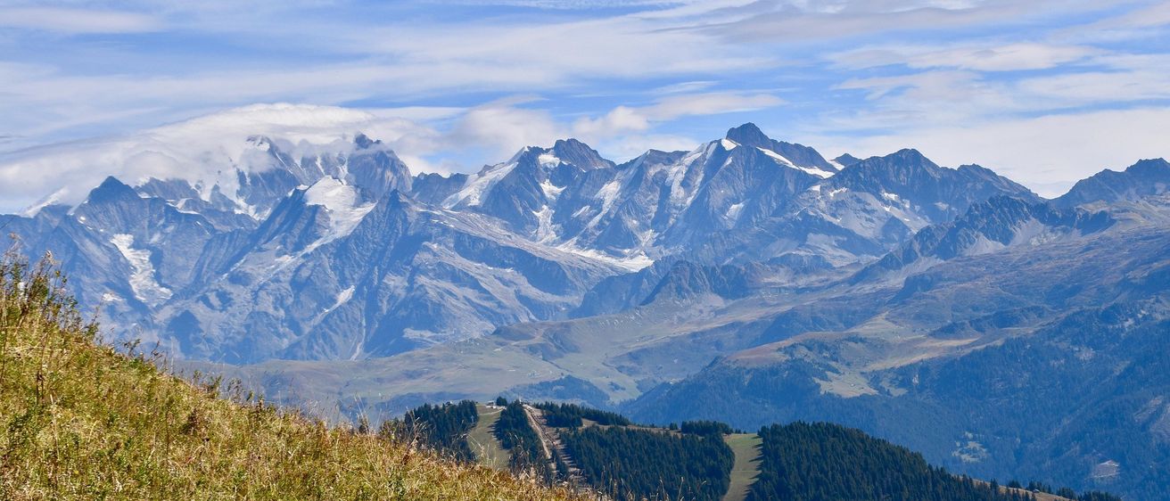 Eine Bergkette mit schneebedeckten Gipfeln, umgeben von grünen Bäumen und Gras. Der Himmel ist blau mit einigen Wolken.