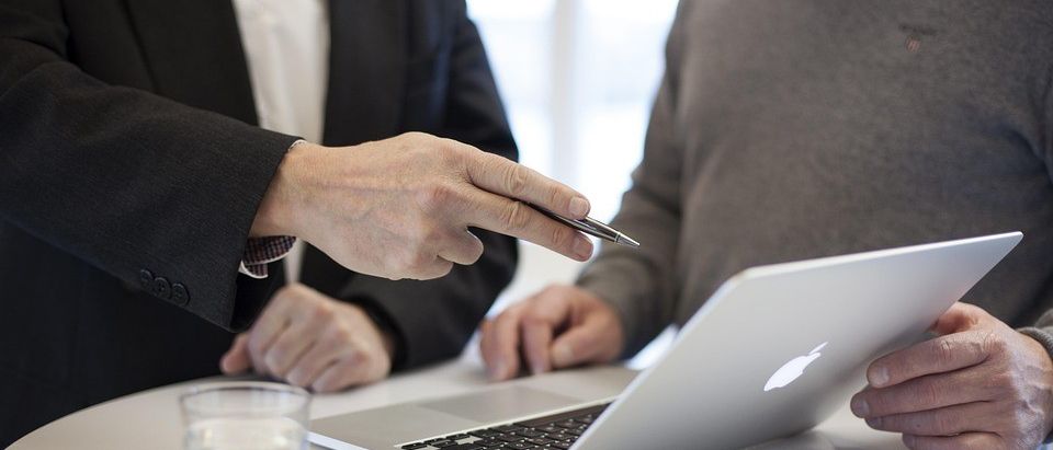 Zwei Personen sitzen an einem Tisch, eine hält einen Stift und zeigt auf einen offenen Laptop. Ein Glas Wasser und Papiere sind auf dem Tisch.