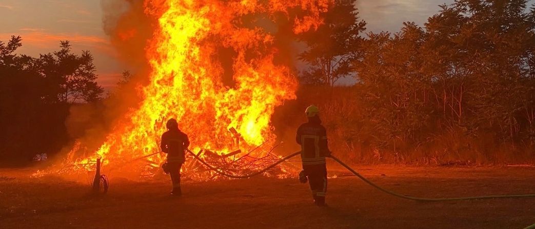 Zwei Feuerwehrleute löschen bei Dämmerung ein großes Feuer auf einem offenen Feld, umgeben von Bäumen.