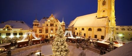 A nighttime view of a festive town square with a church tower, a decorated Christmas tree, and people walking around during the holiday season.