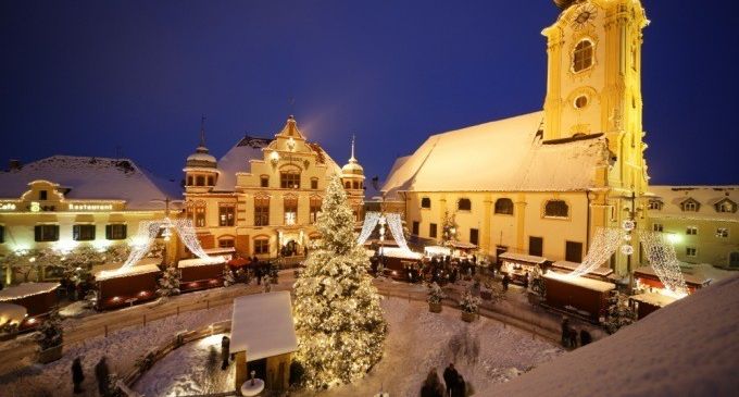 A nighttime view of a festive town square with a church tower, a decorated Christmas tree, and people walking around during the holiday season.